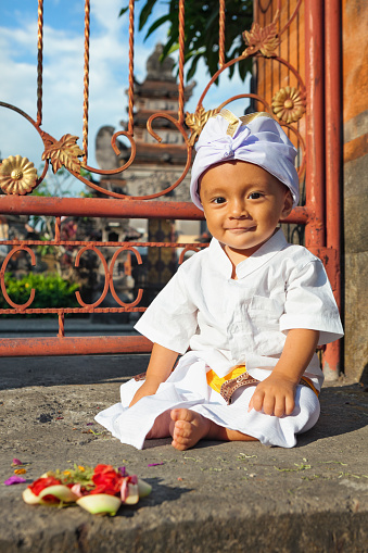 Portrait of balinese baby boy with smiling face in traditional costume Sarong sitting in hindu temple at religious ceremony. Bali island children lifestyle and national culture of Indonesian people.