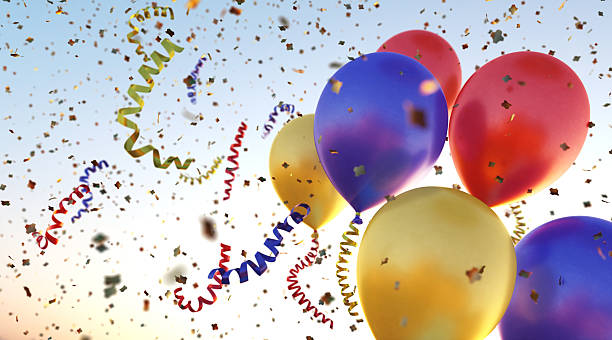 A bundle of metallic colored balloons flying through many colorful confetti flakes and swirled ribbons. The whole scene is situated on a clear sky background.