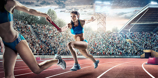 Running Pass on . Stadium A woman athletes sprinting and passing race baton on the track on the . stadium. The bleachers are full of spectators. The sky is blue and cloudy. The woman is wearing an unbranded bra top with small bottoms. relay photos stock pictures, royalty-free photos & images