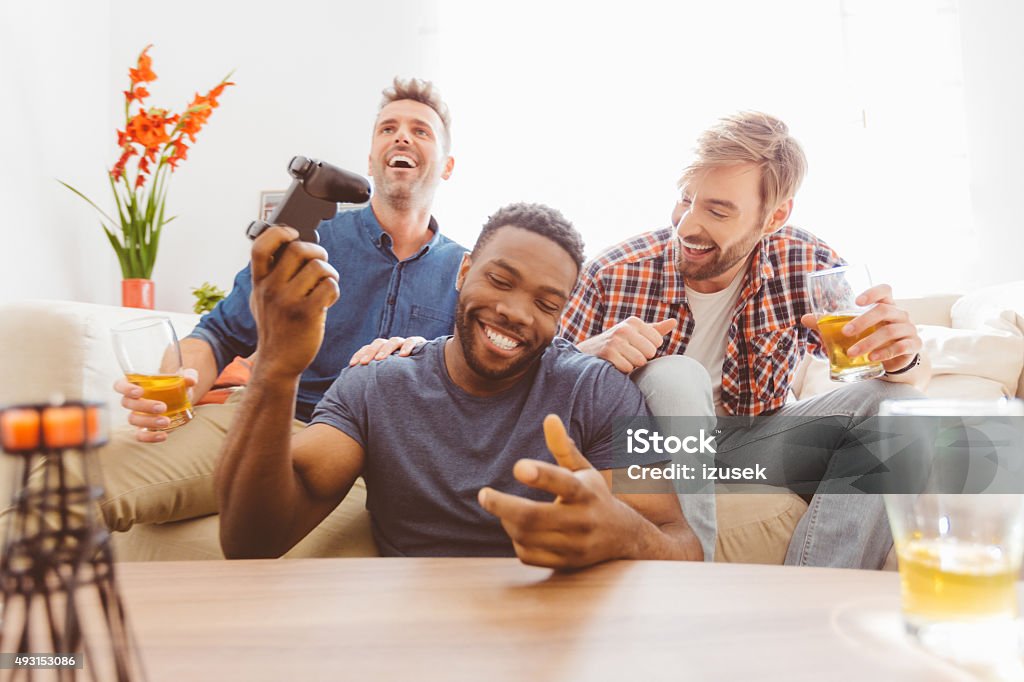 Three guys playing video games Multi ethnic - caucasian and afro american - friends sitting on sofa at home, playing video games and drinking beer.  Control Stock Photo