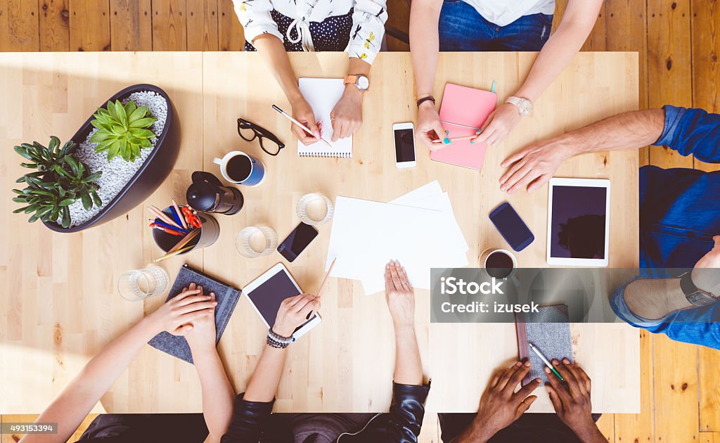 Team brainstorming, high angle view on the table High angle view of group of people sitting at the conference table, discussing, brainstorming. Digital tablets, smart phones, notebooks, coffees on the table. Unrecognizable people. High Angle View Stock Photo