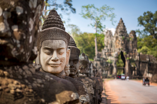 A newly made Buddhist head stands out from the other ancient statues while on the rear, some tuk tuk are driving through the gate to Angkor Thom, another temple complex of the famous Angkor Wat heritage site. Selective focus on the new stone head.