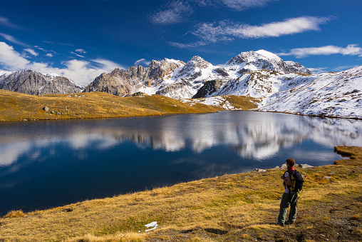 Hiker looking the outstanding view of high altitude blue lake and majestic snowcapped mountain peak in autumn season. Wide angle shot in the Italian French Alps.