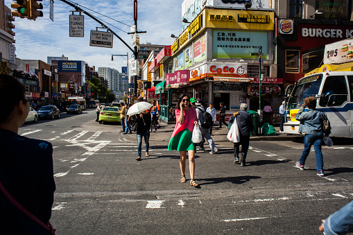 Flushing, NY, USA - September 21, 2015: A woman with a parasol and another woman in a colorful dress talking on her phone, cross Main Street in Flushing Queens' Chinatown. In the background we see other pedestrians, a green taxi cab, a bus at a bus stop, and various businesses, including a CoCo tea and juice shop, Burger King, and a Western Union with Chinese script.