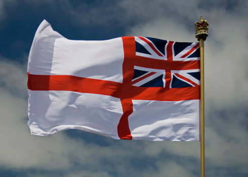 A photo of the Bristish Royal Naval Flag, flying against a blue sky with some light clouds, and a crown on the top of the flag pole