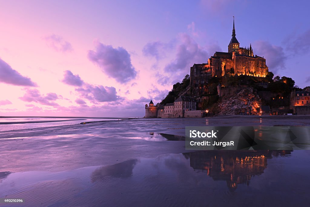 Reflections of Mont Saint-Michel Lit in the Evening The famous island of Mont Saint Michel in Normandy, France, is here seen lit in the early evening, just after sunset. Reflections of the mountain and beautiful colors of the sky can be seen in the low tide water at the bay. This is a purple-toned landscape image and there are no people in it. Mont Saint-Michel Stock Photo
