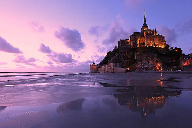The famous island of Mont Saint Michel in Normandy, France, is here seen lit in the early evening, just after sunset. Reflections of the mountain and beautiful colors of the sky can be seen in the low tide water at the bay. This is a purple-toned landscape image and there are no people in it.