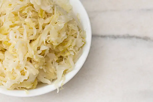 An overhead close up horizontal photograph of a white bowl sitting on a marble counter and containing some fresh, homemade sauerkraut.