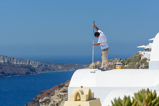 Blond woman with beach hat and turqoise dress visiting the old town of Oia, Greece. Standing on rooftop terrace, hands outstretched and looking at the sea and the volcano island. Ship sailing at the sea.
