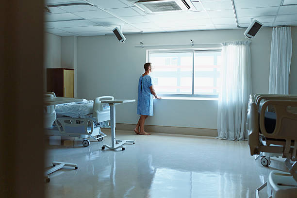 thoughtful patient looking through window - examination gown fotografías e imágenes de stock