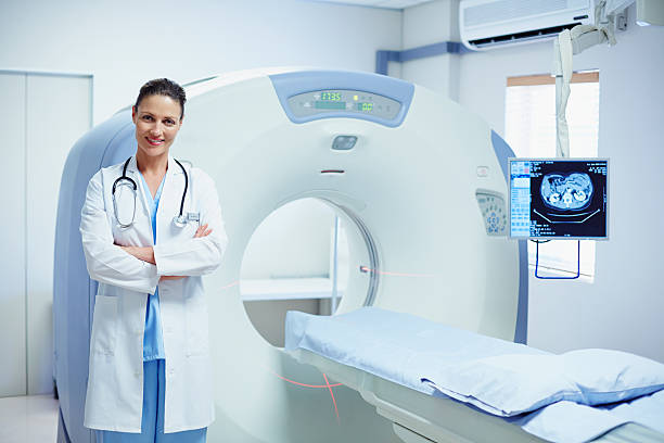 confident female doctor standing by ct scanner - tomografía fotografías e imágenes de stock