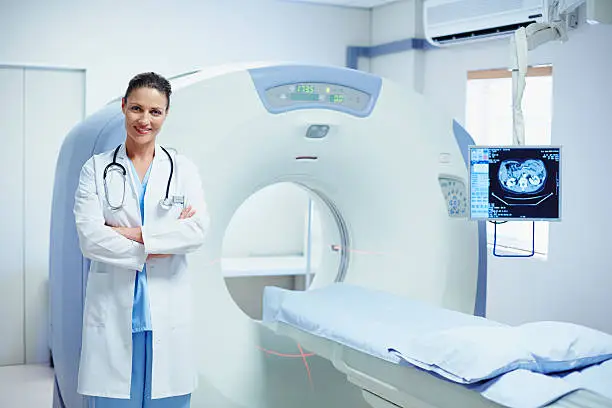 Portrait of confident female doctor standing arms crossed by CT scanner in hospital