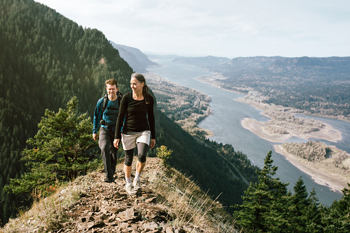 A fit older adult couple in their 50's hike up a rocky trail on a mountain ridge, the beautiful Columbia river gorge spreading out behind them.  They smile as they continue up the ascent.  Horizontal with copy space.