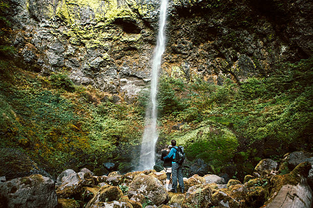 A fit older adult couple in their 50's pause from their hike to view Elowah falls in the Columbia river gorge.  They stand at the rock base of the waterfall, their arms around each other while taking in the scenery.  Horizontal image with copy space.