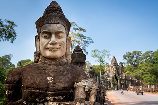 A newly made Buddhist head stands out from the other ancient statues while on the rear, some tuk tuk are driving through the gate to Angkor Thom, another temple complex of the famous Angkor Wat heritage site. Selective focus on the new stone head.