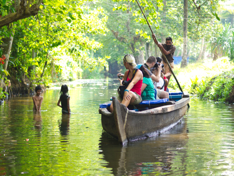 Cochin, India - December 21, 2013: Tourists having ridden on traditional Kerala boat with boat driver in cannel surrounded with vegetation. Two Indian kids are playing in water, while on water is strong sun reflection coming out from background.