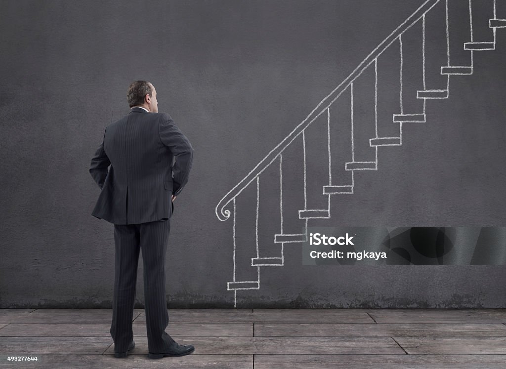 Businessman with Staircase Sketched on the Wall Rear view of businessman standing on wooden floor with staircase and banister sketched (chalk drawing) on the wall. 2015 Stock Photo