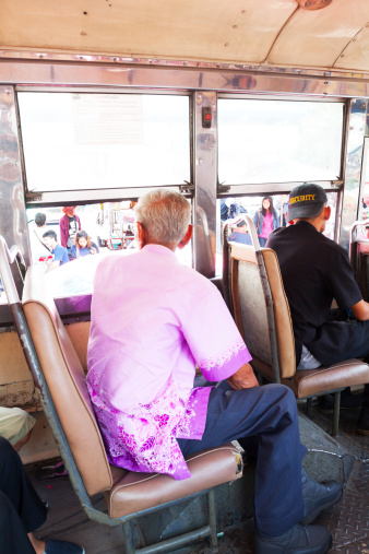 Bangkok, Thailand - January 28, 2014: Some people are sitting inside of bus in Bangkok and are watching to sidewalk and people outside. There are several groups of Thai people. On seats is Thai senior and a younger Thai man. Younger man is wearing a cap.