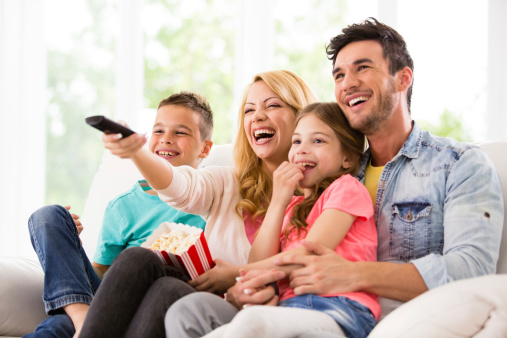 A happy family watches TV at home.  A dad in a denim shirt holds a little girl in a pink t-shirt.  A smiling mom holds a container of popcorn and directs a remote control toward a television.  A young boy wears a turquoise t-shirt and a pair of blue jeans.