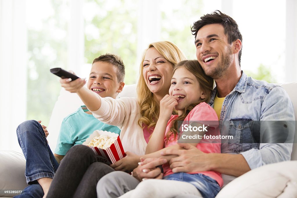Happy family watching TV and eating popcorn A happy family watches TV at home.  A dad in a denim shirt holds a little girl in a pink t-shirt.  A smiling mom holds a container of popcorn and directs a remote control toward a television.  A young boy wears a turquoise t-shirt and a pair of blue jeans. Family Stock Photo