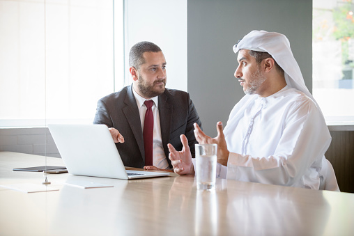 Mid adult ethnic Arab businessman with mature male colleague wearing traditional clothing. Two male adult men using laptop. Mature Arabic businessman wearing Thobe or Kandura and Ghoutra headdress explaining.