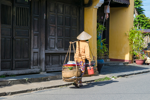 Hoi An, Vietnam  - September 7, 2014: Woman carrying the heavy yoke loaded with food to sell on the street of Hoi An on September 7, 2014