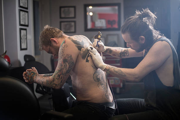 Male artist tattooing man's back A photo of male artist tattooing man's back. Tattoo expert is working in studio. Shirtless male customer getting his back tattooed. body adornment rear view young men men stock pictures, royalty-free photos & images