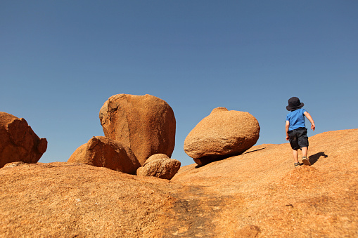 Little boy is climbing a big granite rock in Spitzkoppe in Namibia, Southern Africa. The boy is here seen from the back, almost on top of the hill with only rocks around and and blue sky in front of him. This image of a young traveller exploring nature could fit various concepts related to discovery, adventure, travelling with children, etc. as the location is rather generic.