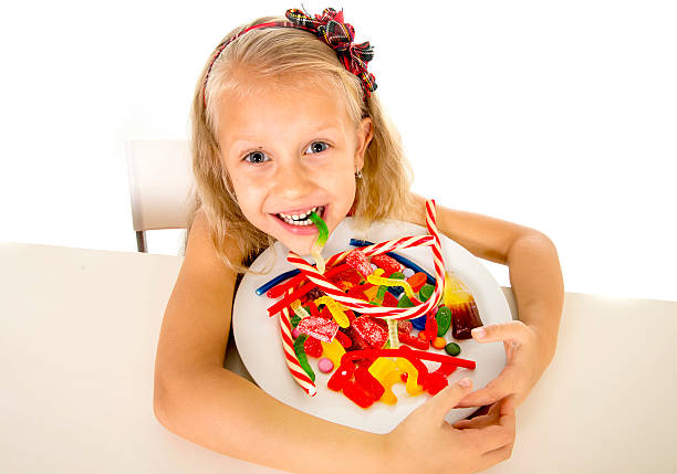 pretty happy child eating candy sweet sugar abuse dangerous diet pretty happy Caucasian female child eating dish full of candy holding the dish in sweet sugar abuse dangerous diet and unhealthy nutrition concept isolated on white background eye candy stock pictures, royalty-free photos & images