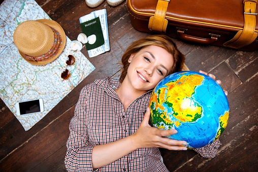 Top view photo of beautiful blonde girl lying on wooden floor. Young woman smiling, holding globe and looking at camera. Passport, tickets, mobile phone, hat, suitcase and map are on floor