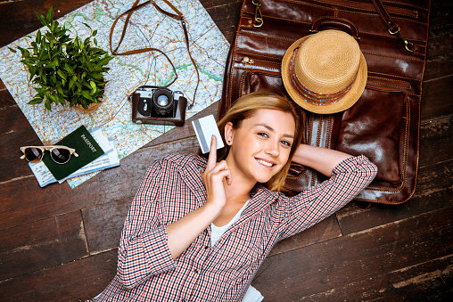 Top view photo of beautiful blonde girl lying on wooden floor. Young woman smiling, holding credit card and looking at camera. Passport, tickets, vintage camera, hat and map are on floor