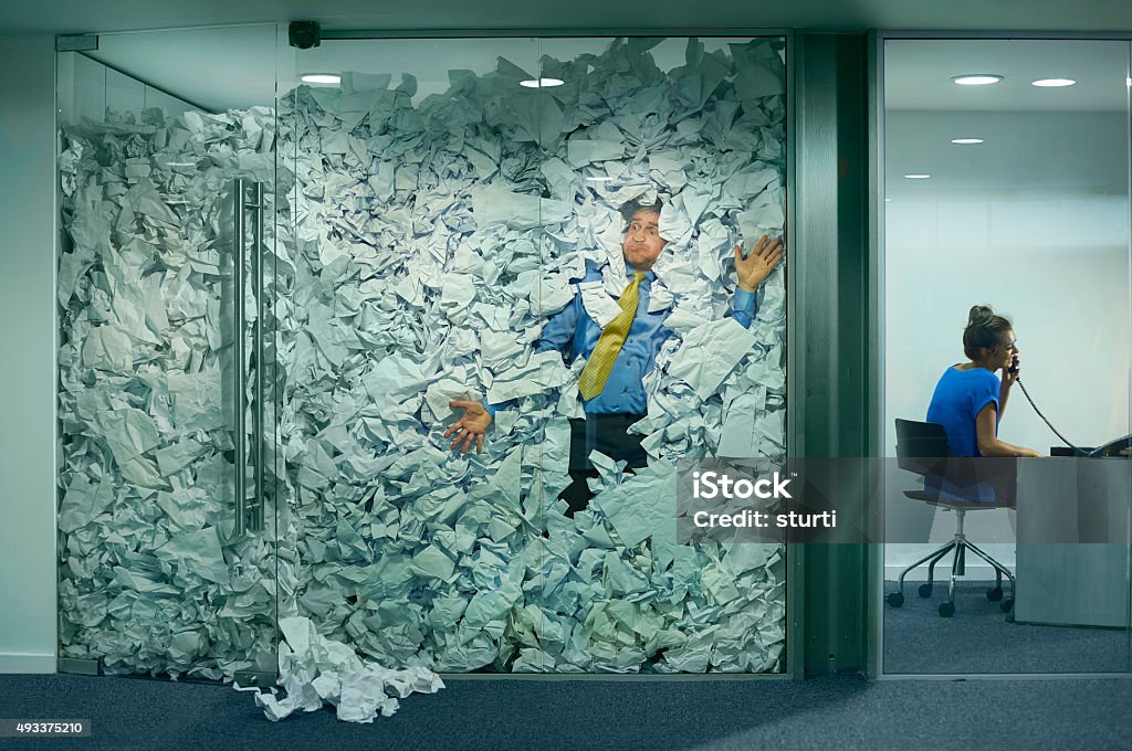 stuck at the office A businessman is trapped in his glass office by a surplus of discarded ideas on paper . His colleague in the next office is working more efficiently and is oblivious to him being trapped . Office Stock Photo
