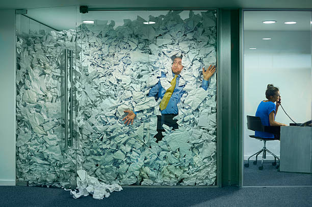 stuck at the office A businessman is trapped in his glass office by a surplus of discarded ideas on paper . His colleague in the next office is working more efficiently and is oblivious to him being trapped . bureaucracy stock pictures, royalty-free photos & images
