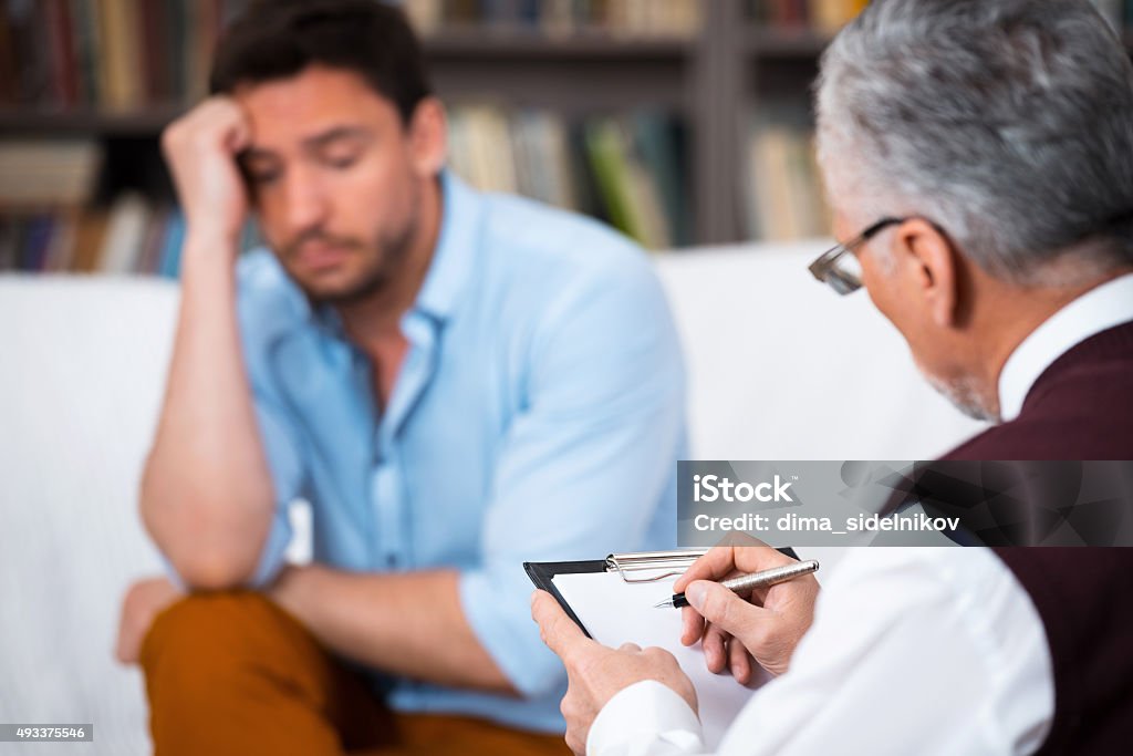 Concept for consultation with psychologist Sad young man talking with psychologist. Psychologist taking notes. There are many books in psychologist office Mental Health Professional Stock Photo