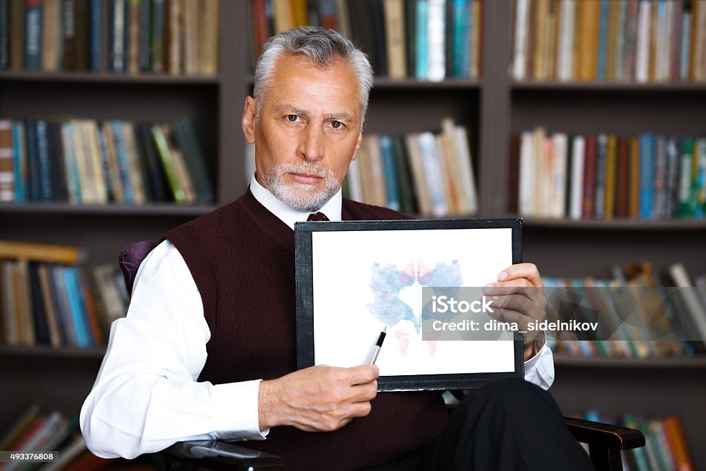 Concept for consultation with psychologist Portrait of male psychologist. Psychologist standing near big cupboard with books in psychologist office and holding Rorschach paintings Rorschach Test Stock Photo