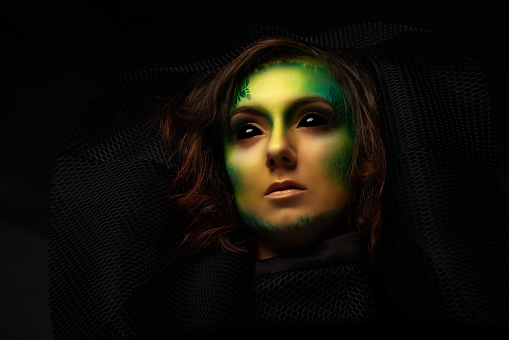 portrait of Halloween demon woman with face paint, green makeup looking up with misterious look, black background.