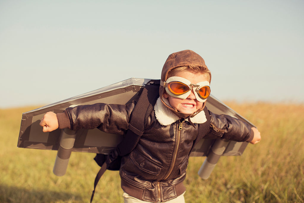 A young boy wearing a bomber jacket and goggles is ready to fly his homemade jetpack. He is standing in the tall grass with arms raised on a hill in England ready for adventure.