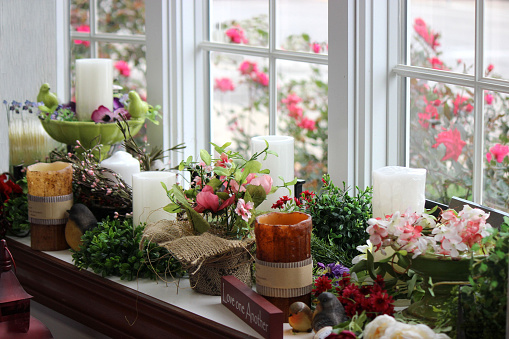 Beautiful photo of candles, candlestick holders, flowers, vases, wreaths, plants, birds and gifts on a window seat in front of a bay window in a gift shop.  Bright pink or fuchsia coloured roses can be seen in the garden outside the window.  The caption \