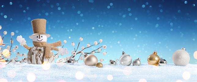 Happy snowman holds a bag with Christmas presents, dressed in warm clothes, mittens, hat and scarf in a frosty winter day. Winter or Christmas background or banner with defocused lights and lens flare.