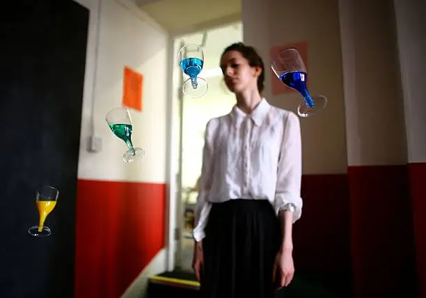 Young woman in white blouse standing in front of four floating wine glasses, each filled with different colored drinks.