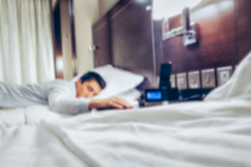 Out of focus background of businessman sleeping on bed and his hand front of the alarm clock at 6:30 AM, Wake up early morning concept purposely with blurred effect