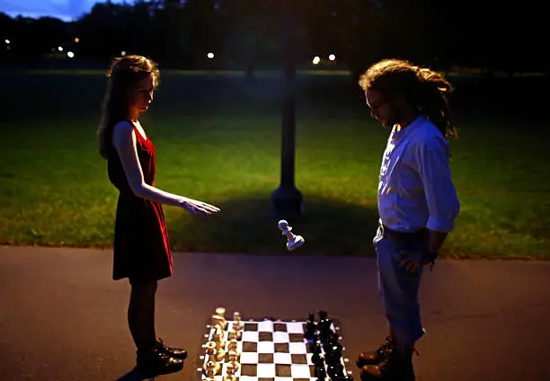 Woman in red velvet dress and man with dreadlocks playing chess in a dark park at night as the woman makes a pawn levitate off the ground.
