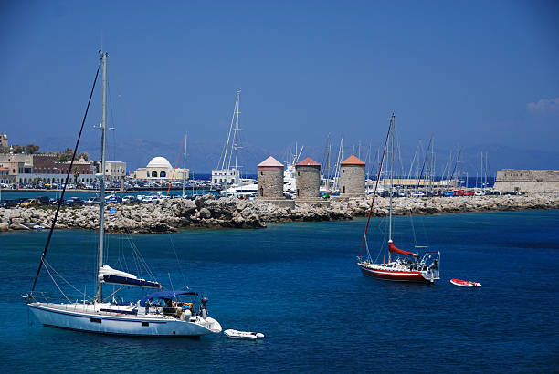 Sailboats anchored in the harbor of Old Town, Rhodes with iconic Greek windmills in the background.