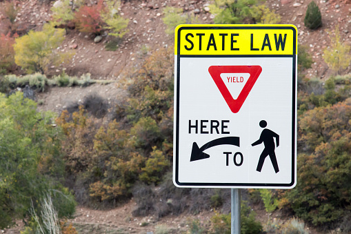 Traffic sign that reminds motorists of the state law to yield to pedestrians.  The sign is against a bush covered hillside in Colorado.  The sign has a yield sign, an arrow, and a symbol of a person walking.