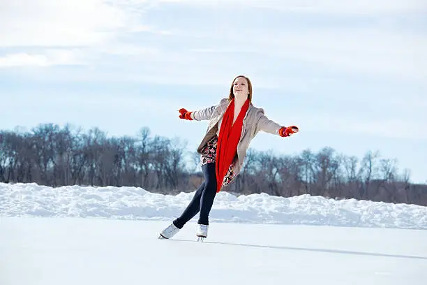 A cute teenage girl leisurely figure skating outdoors on a pond ice rink on a cold winter day. She wears a gray jacket, skirt, tights, red neck scarf and mittens for warmth as she extends arms and glides on one foot in a Lake Harriet urban park, Minneapolis, Minnesota.