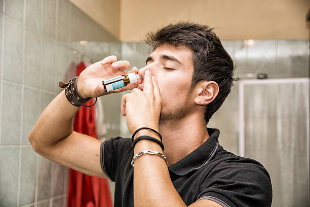 Young Man Sniffing Nose Spray in Bathroom Head and Shoulders Close Up of Young Attractive Man with Dark Hair Sniffing Nose Spray with Eyes Closed in Home Bathroom nasal spray stock pictures, royalty-free photos & images