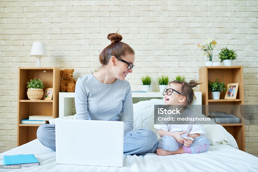 Joyful social media users Mother with laptop and little daughter with smart phone sitting on bed, looking at each other and laughing Child Stock Photo