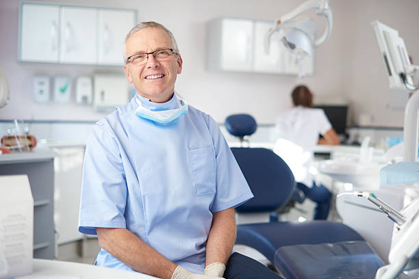 mature male dentist portrait A mature male dentist is looking proudly to camera from his dentist's office. In the background a dental nurse is prepping for the next patient. dentist photos stock pictures, royalty-free photos & images