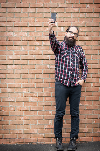 A bearded hipster guy taking a selfie as he stands by a brick wall.
