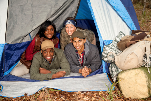 Diverse group of young adults gather in tent ready to enjoy camping trip in forest. Camping gear stacked outside tent. 